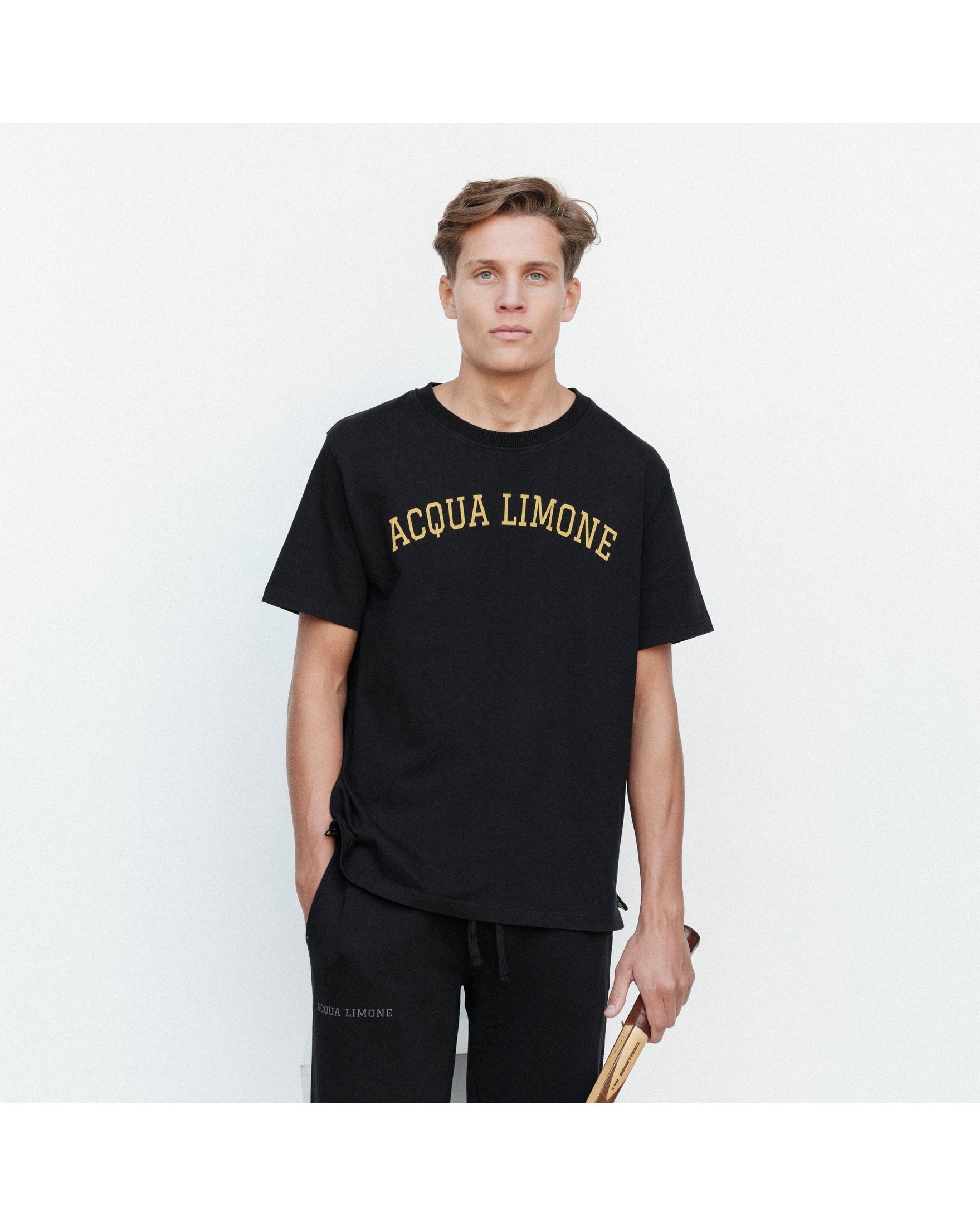 Man wearing a black 'Acqua Limone' t-shirt and holding a tennis racket against a white background