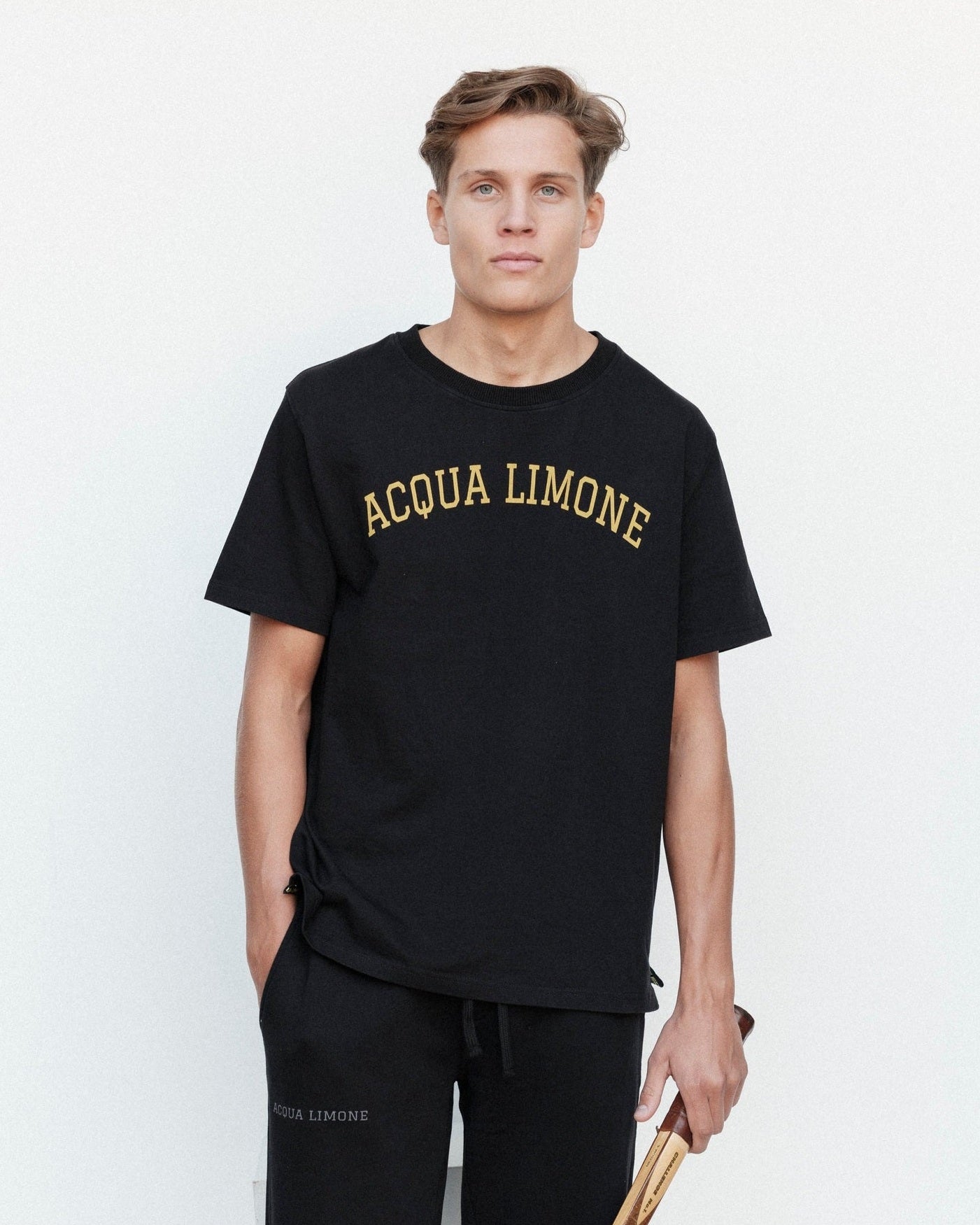 Man wearing a black 'Acqua Limone' t-shirt and holding a tennis racket against a white background