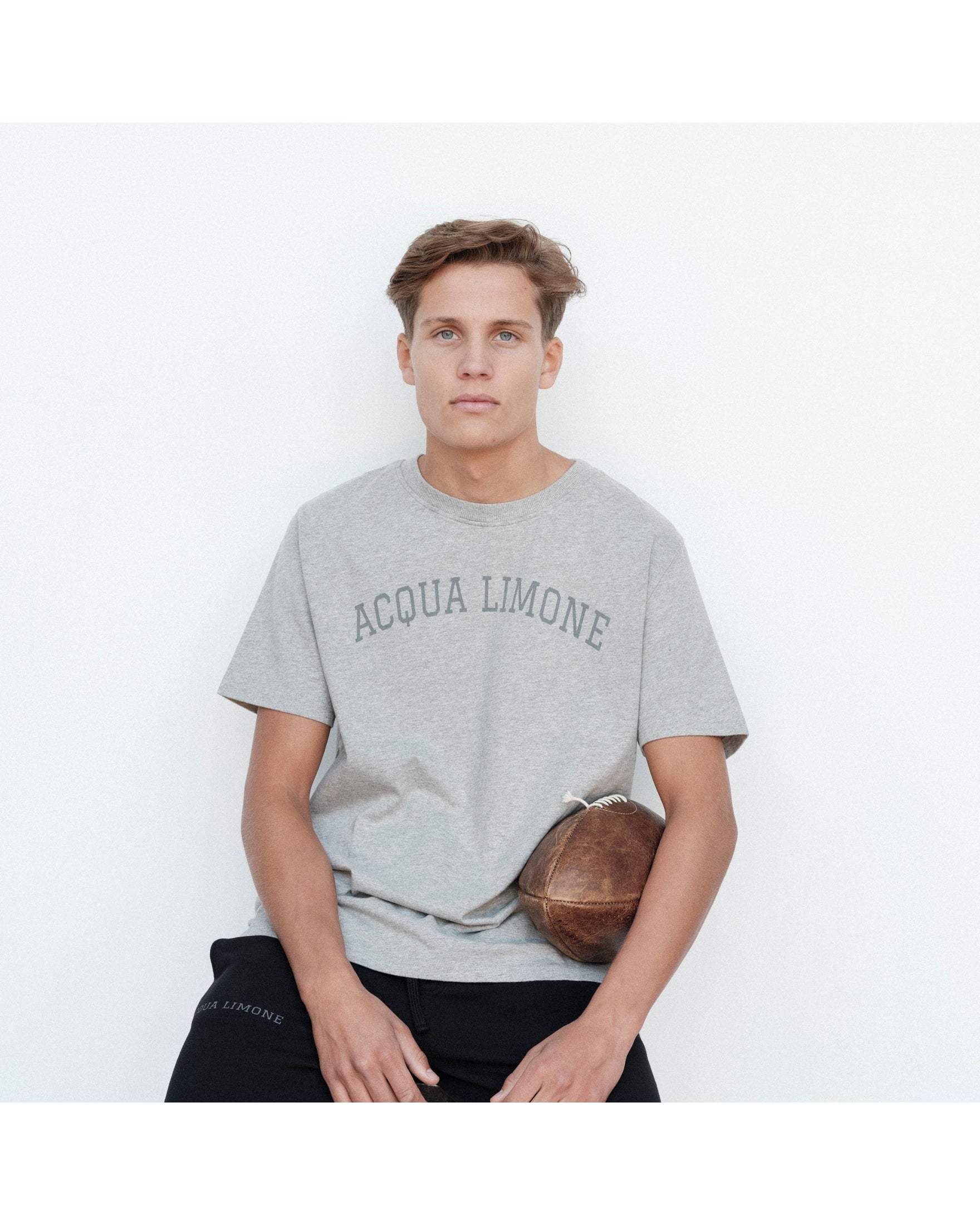 Person wearing a gray 'Acqua Limone' t-shirt holding a football against a white background