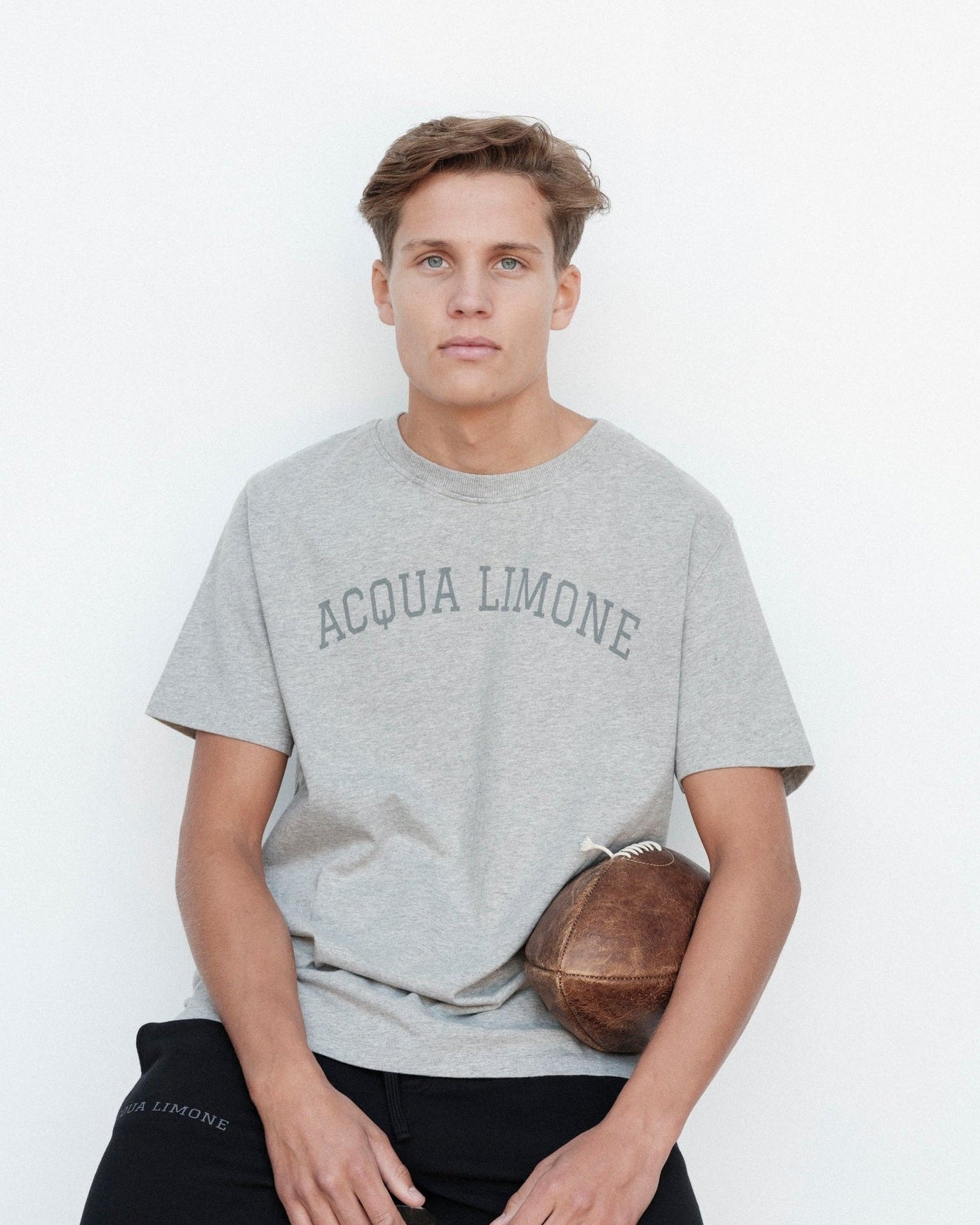 Person wearing a gray 'Acqua Limone' t-shirt holding a football against a white background