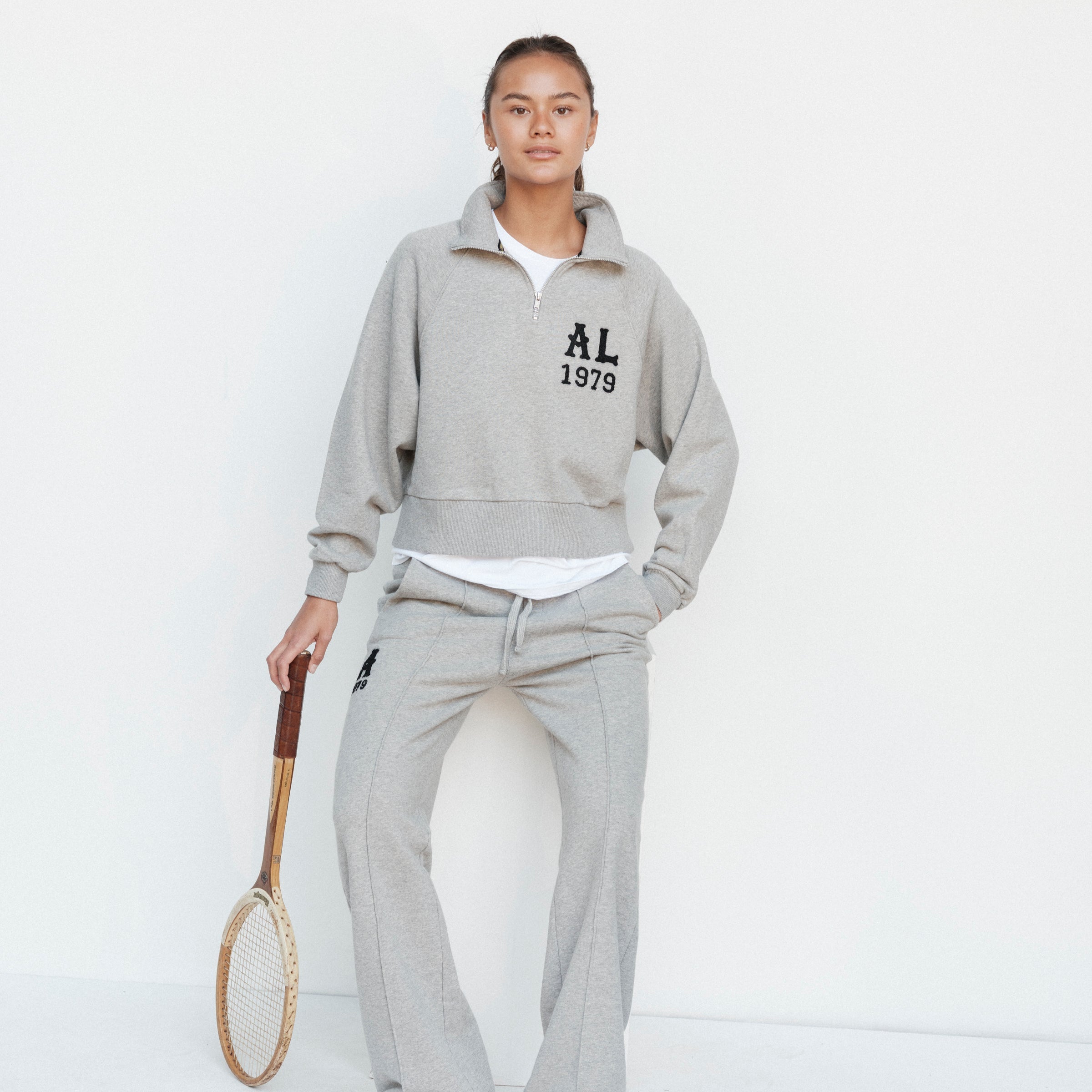 Person wearing a gray tracksuit with 'AL 1979' text, holding a tennis racket on a white background