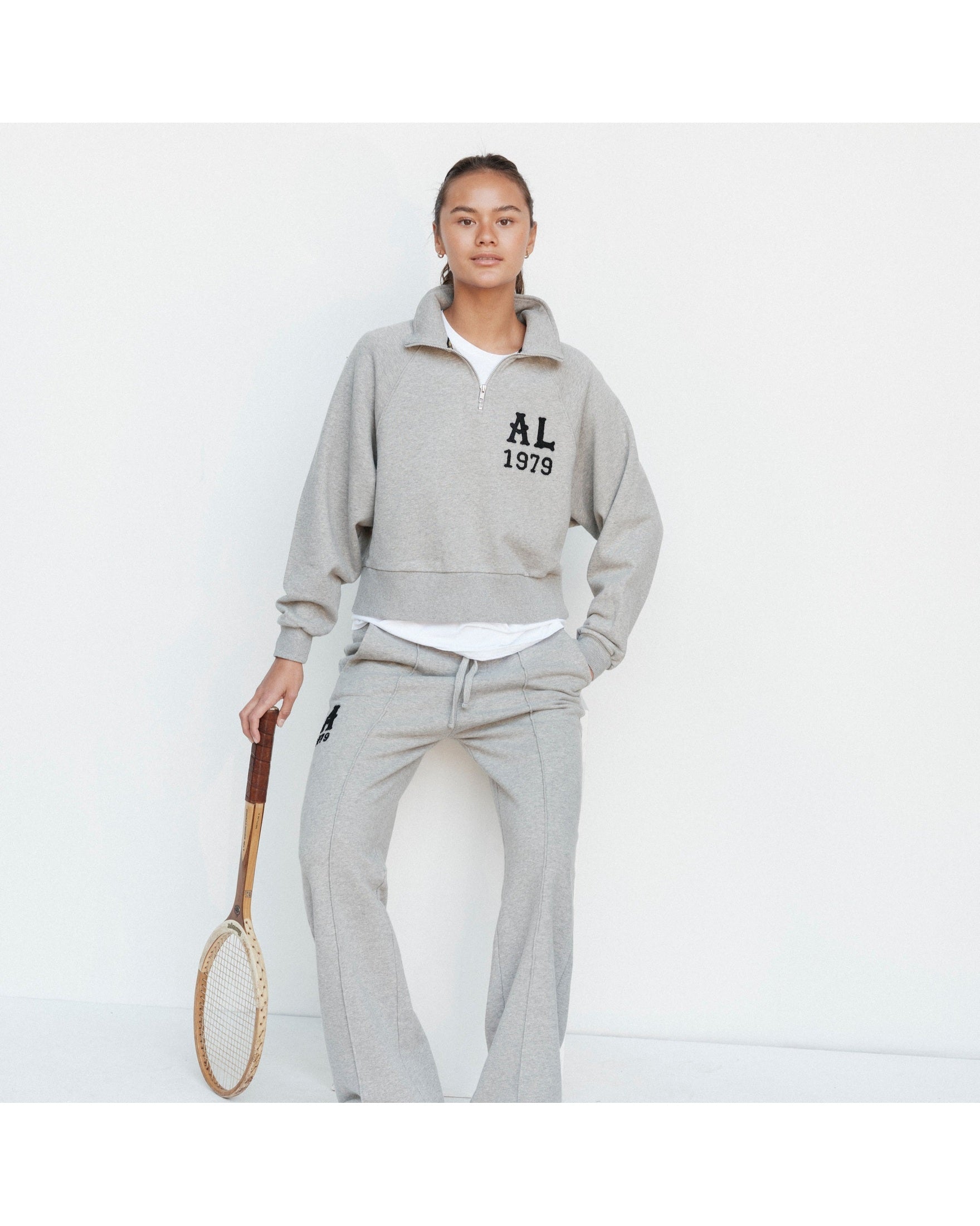 Person wearing a gray tracksuit with 'AL 1979' text, holding a tennis racket on a white background