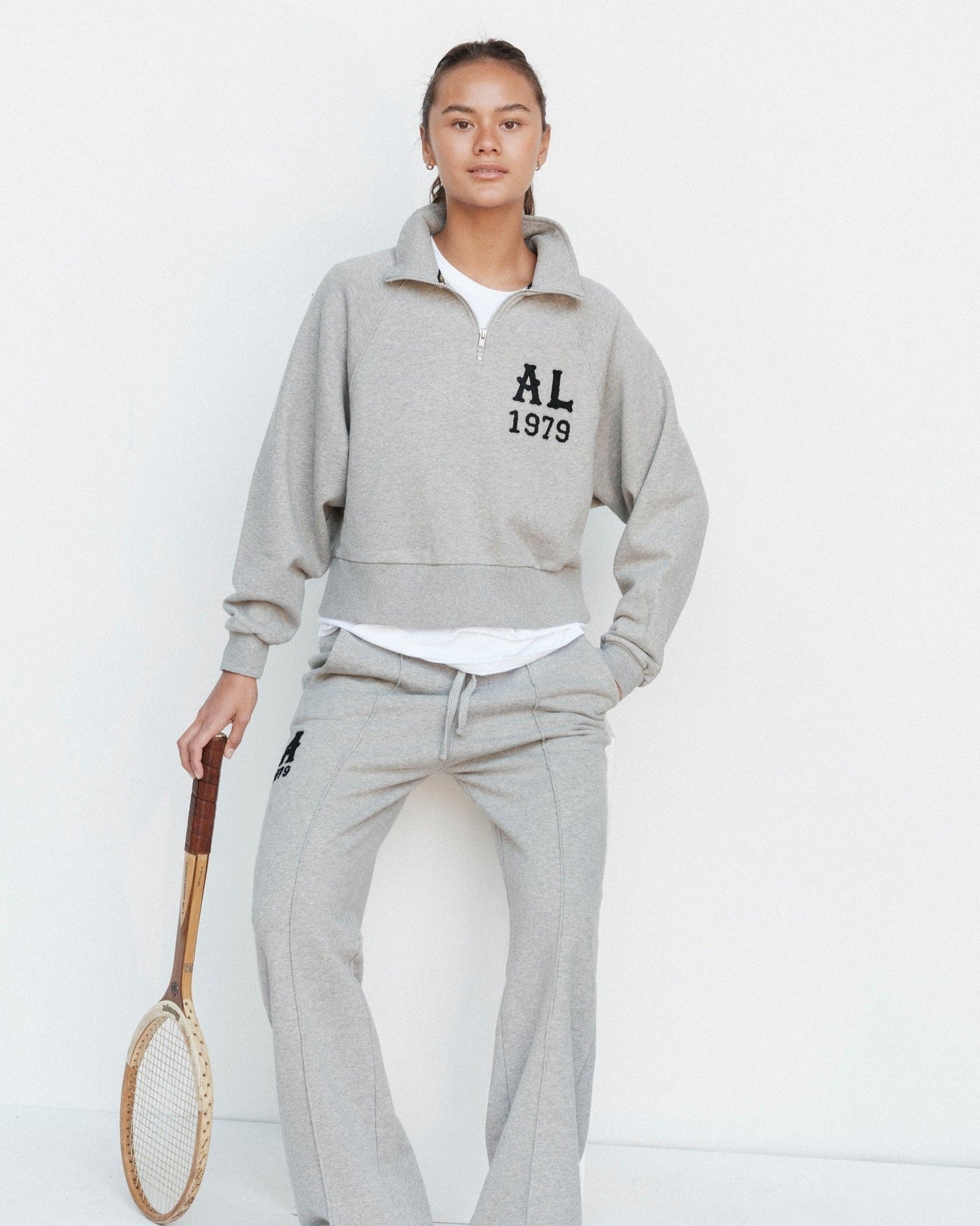 Person wearing a gray tracksuit with 'AL 1979' text, holding a tennis racket on a white background