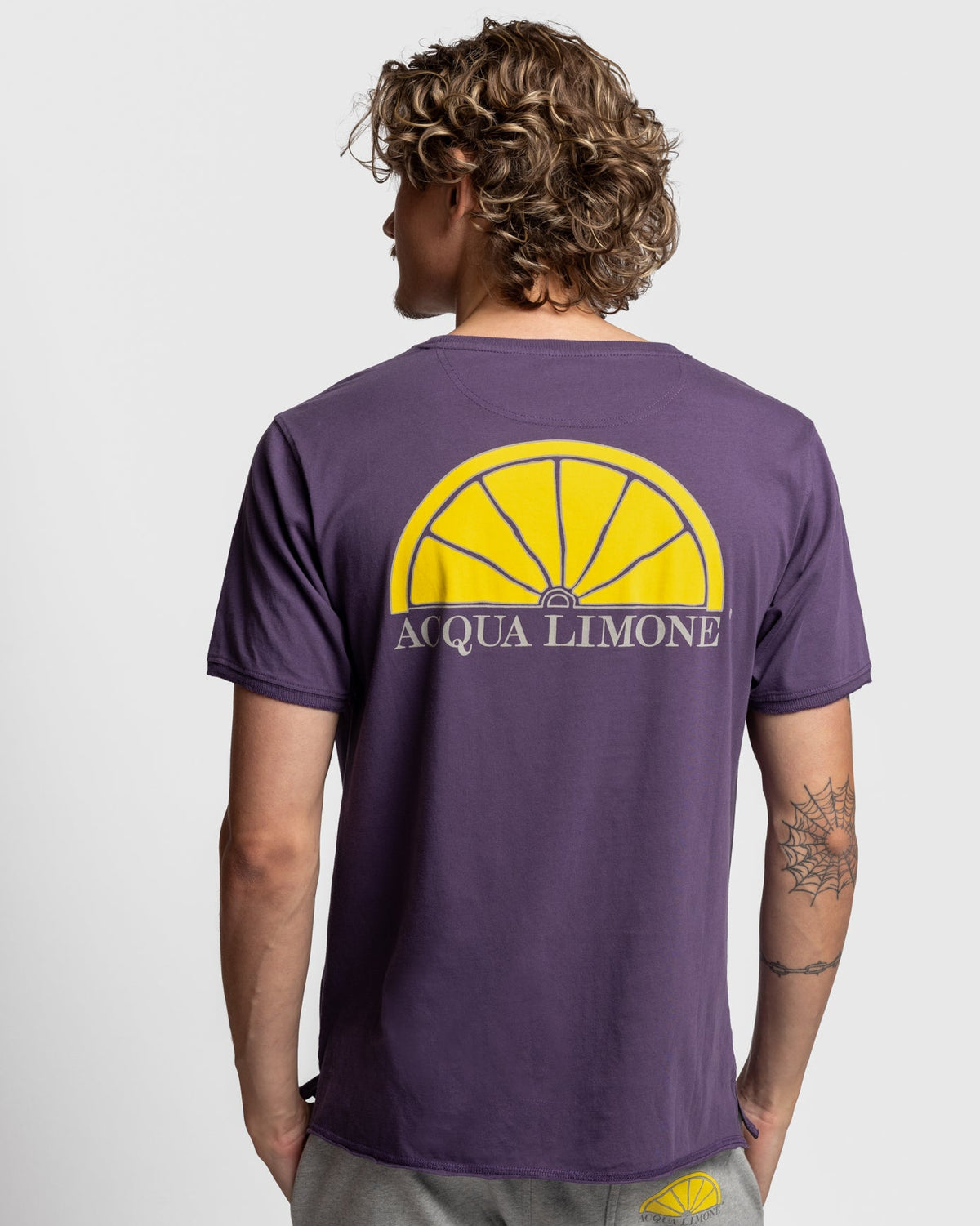 A man viewed from behind wearing a purple t-shirt with a large yellow lemon graphic and the text 'ACQUA LIMONE' printed on the back, paired with gray sweatpants against a light gray background.
