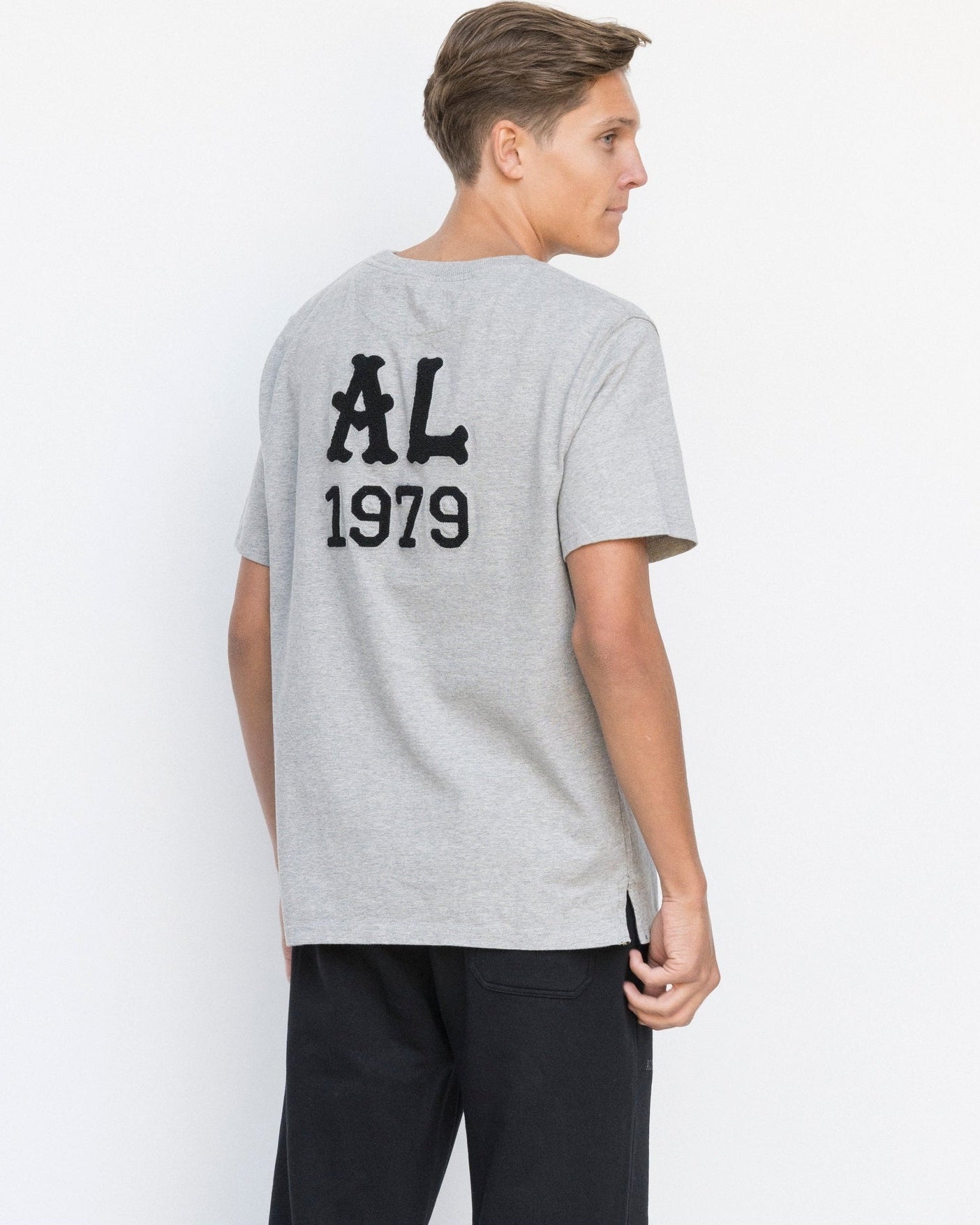 Person wearing a gray t-shirt with 'AL 1979' text on the back, standing against a white background.