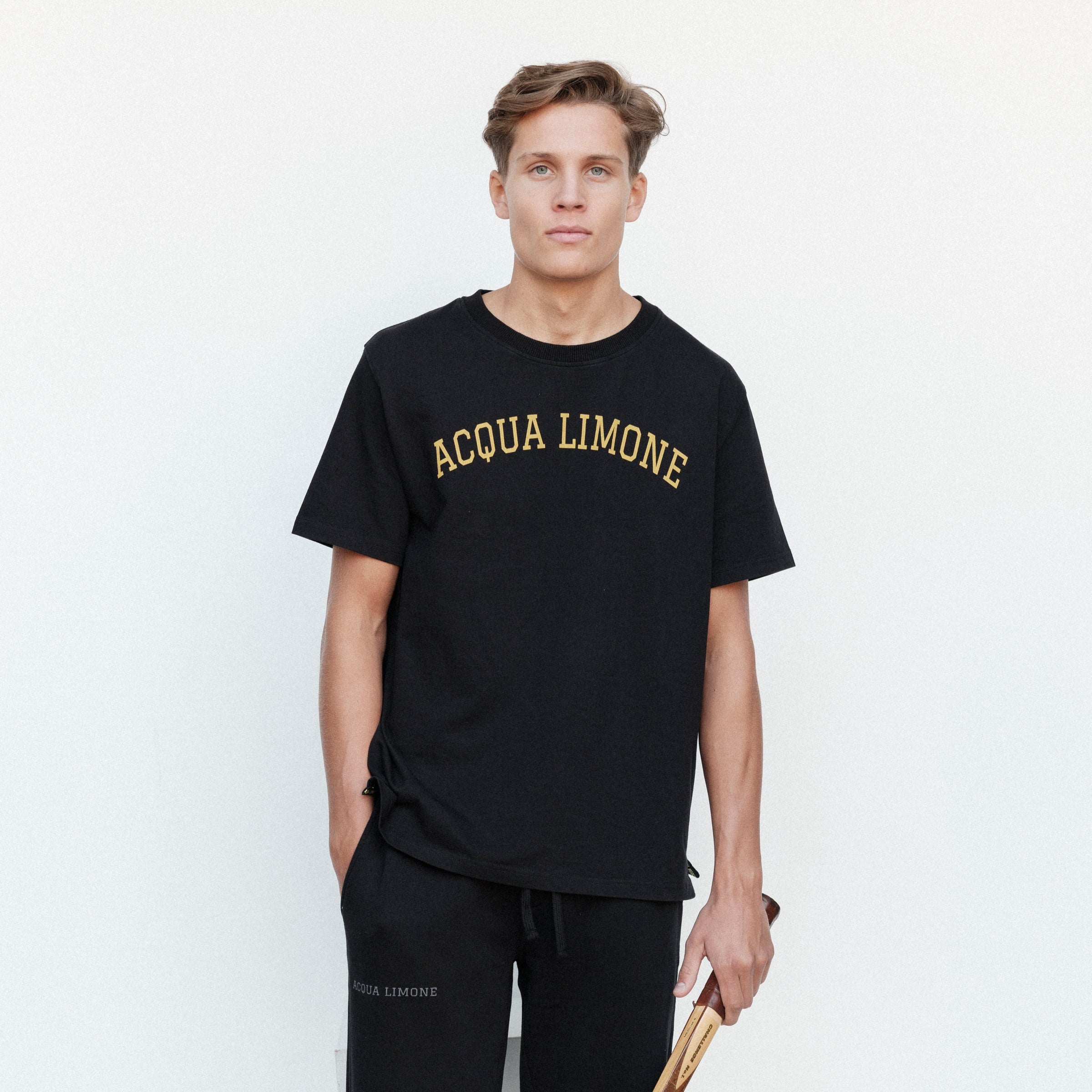 Man wearing a black 'Acqua Limone' t-shirt and holding a tennis racket against a white background