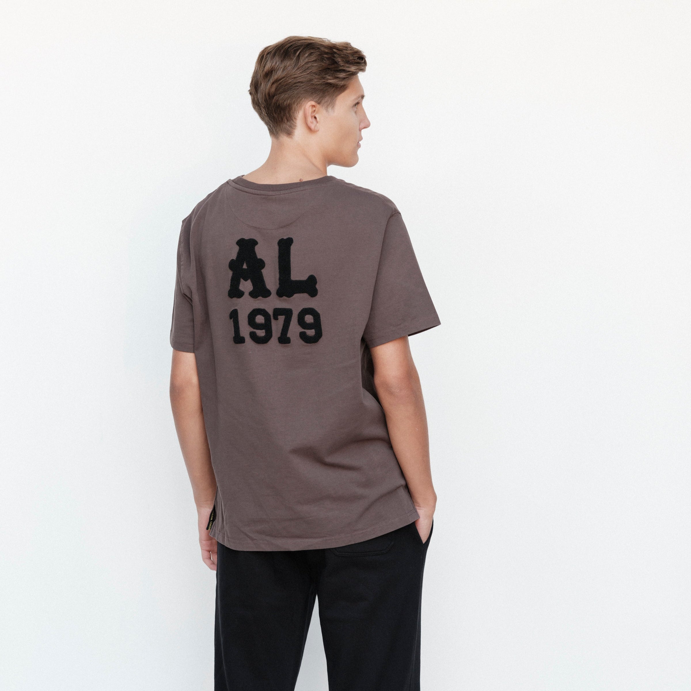 Person wearing a brown t-shirt with 'AL 1979' on the back against a white background