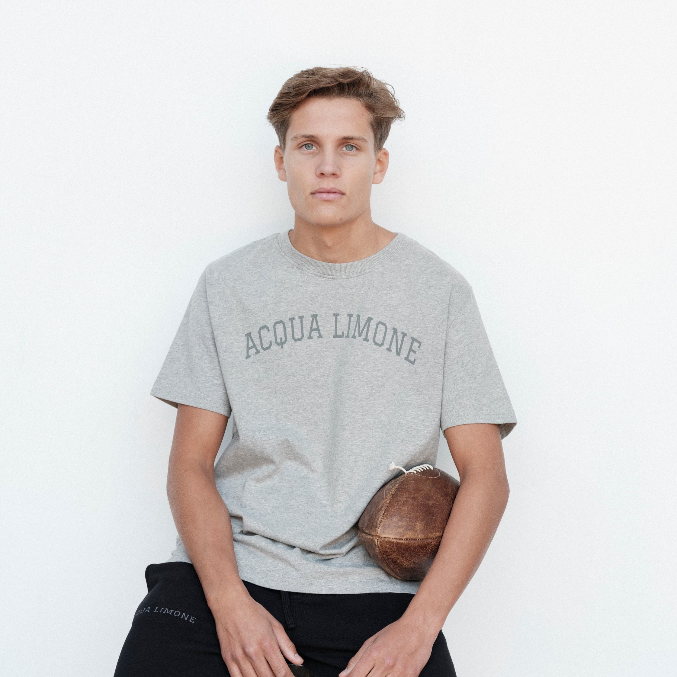 Person wearing a gray 'Acqua Limone' t-shirt holding a football against a white background