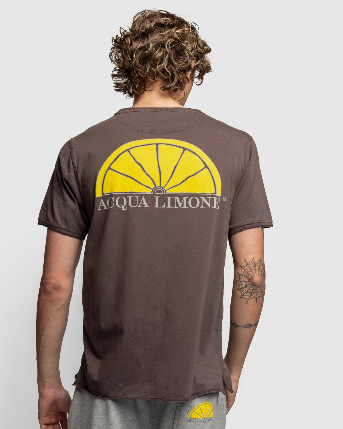 A man viewed from behind wearing a brown t-shirt with a large yellow lemon graphic and 'ACQUA LIMONE' text on the back, paired with gray sweatpants against a light gray background.