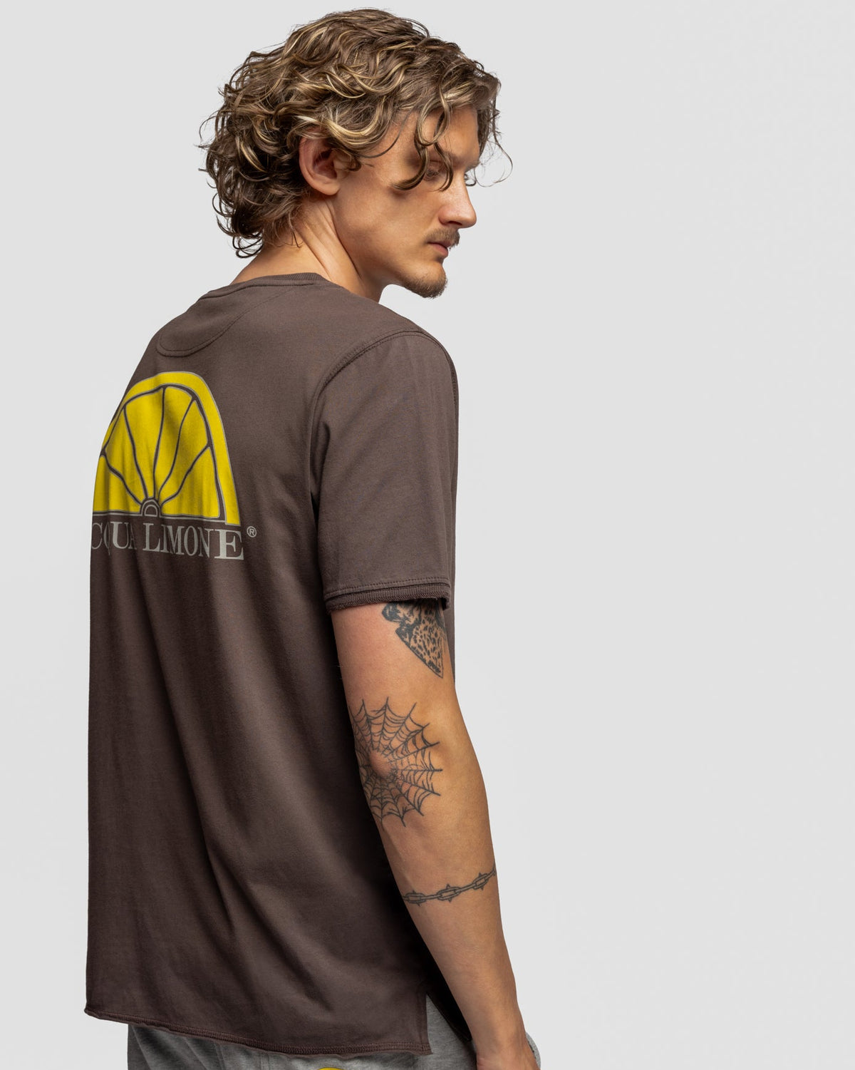 A man wearing a brown t-shirt with a yellow and black bicycle wheel graphic on the back, paired with gray sweatpants, shown from a side-back angle against a light gray background.
