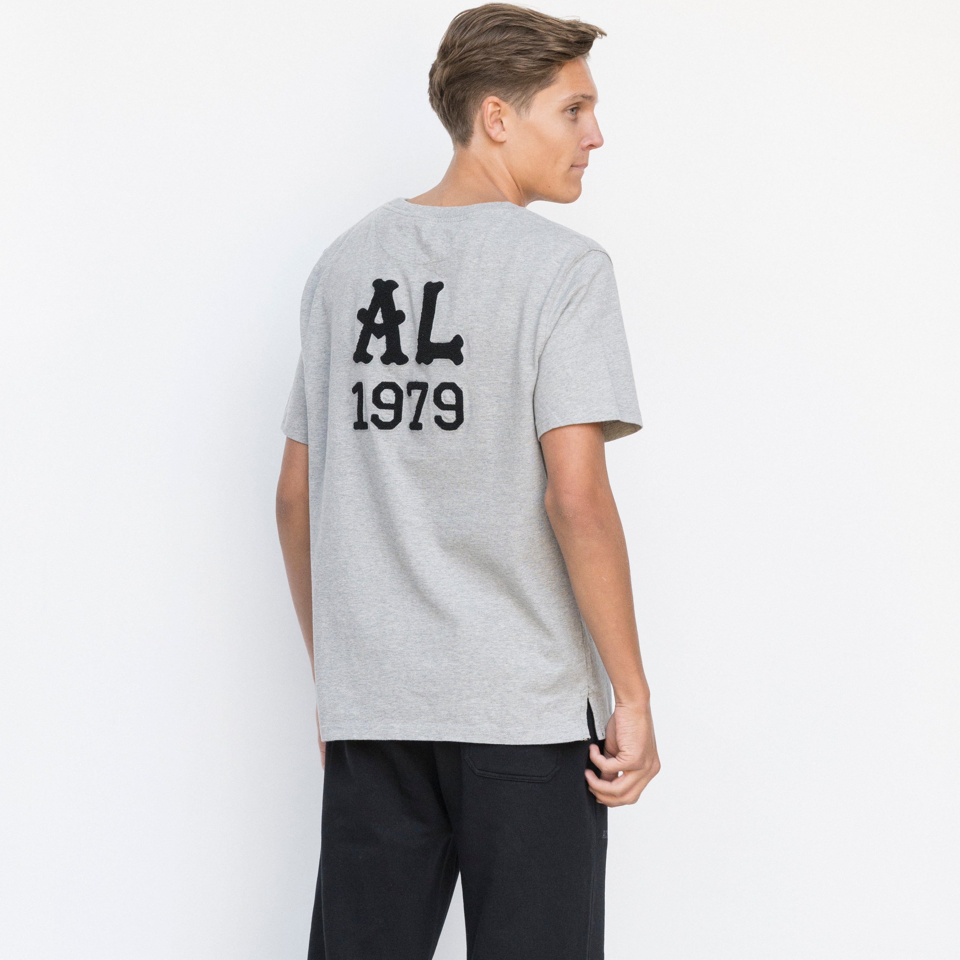 Person wearing a gray t-shirt with 'AL 1979' text on the back, standing against a white background.