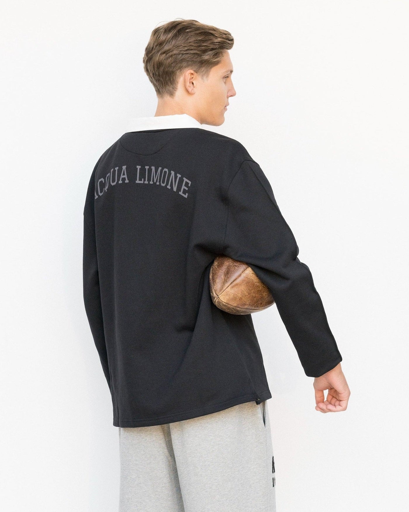 Person wearing a black sweater with 'Acqua Limone' text, holding a football, on a white background